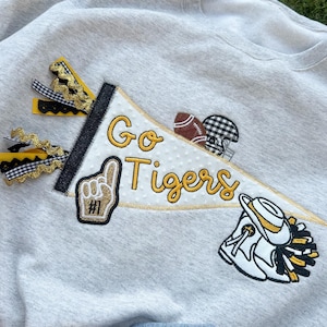 Peut inclure: Un sweat-shirt gris clair avec une bannière décorative qui dit "Go Tigers" en écriture jaune. La bannière présente des écussons sur le thème du football, dont un ballon, un casque et un symbole de main n°1. Des rubans noirs et dorés sont attachés à la bannière.