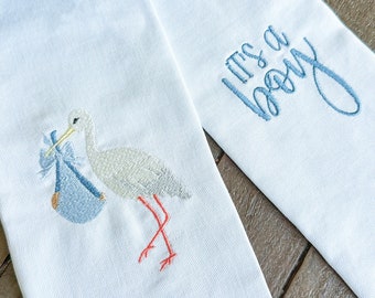 May include: Two white fabric baby shower decorations with blue embroidery. One decoration features a stork carrying a baby in a blue basket. The other decoration says "It's a Boy".