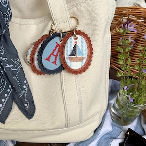 K&ouml;nnte beinhalten: Cremefarbene Tragetasche mit dekorativen Schl&uuml;sselanh&auml;ngern. Die Schl&uuml;sselanh&auml;nger zeigen gestickte Designs eines Segelboots, den Buchstaben "A" und andere nautische Muster. Jeder Schl&uuml;sselanh&auml;nger hat einen gewellten Rand und einen braunen Lederimitat-Rand.
