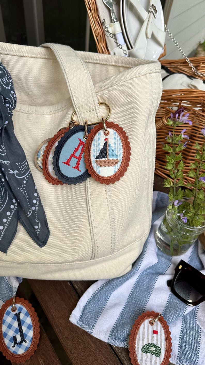 K&ouml;nnte beinhalten: Cremefarbene Tragetasche mit dekorativen Schl&uuml;sselanh&auml;ngern. Die Schl&uuml;sselanh&auml;nger zeigen gestickte Designs eines Segelboots, den Buchstaben "A" und andere nautische Muster. Jeder Schl&uuml;sselanh&auml;nger hat einen gewellten Rand und einen braunen Lederimitat-Rand.