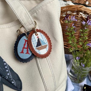 K&ouml;nnte beinhalten: Beige Canvas-Tragetasche mit zwei Schl&uuml;sselanh&auml;ngern. Ein Schl&uuml;sselanh&auml;nger hat ein blaues Oval mit dem roten Buchstaben "A". Der andere Schl&uuml;sselanh&auml;nger hat ein braunes Oval mit einem Segelboot-Design. Ein Picknickkorb und Blumen sind im Hintergrund.