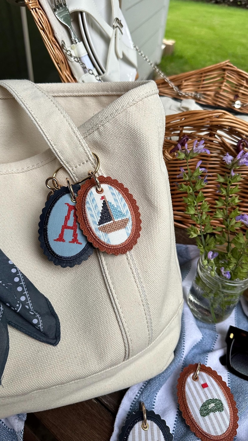 K&ouml;nnte beinhalten: Beige Canvas-Tragetasche mit zwei Schl&uuml;sselanh&auml;ngern. Ein Schl&uuml;sselanh&auml;nger hat ein blaues Oval mit dem roten Buchstaben "A". Der andere Schl&uuml;sselanh&auml;nger hat ein braunes Oval mit einem Segelboot-Design. Ein Picknickkorb und Blumen sind im Hintergrund.