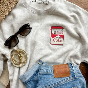Op de afbeelding: Een lichtgrijs sweatshirt met een Diet Coke-afbeelding, gecombineerd met een blauwe spijkerbroek en een zwarte zonnebril. Een gouden armband en een rieten mand zijn ook zichtbaar.