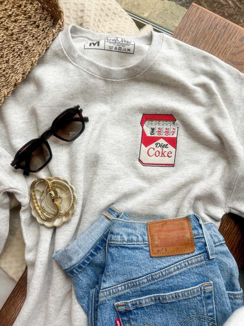 Op de afbeelding: Een lichtgrijs sweatshirt met een Diet Coke-afbeelding, gecombineerd met een blauwe spijkerbroek en een zwarte zonnebril. Een gouden armband en een rieten mand zijn ook zichtbaar.
