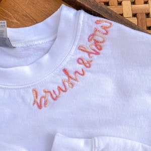 May include: White sweatshirt with the word "brushed" embroidered in pink and orange on the neckline.