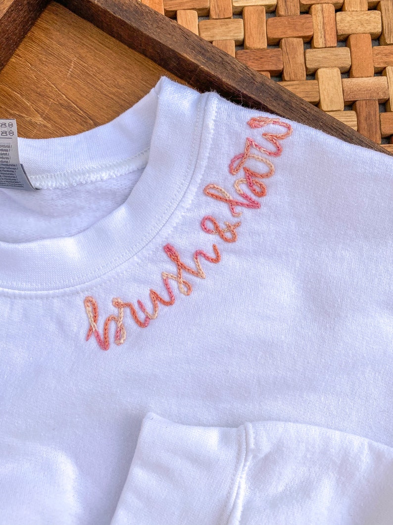 May include: White sweatshirt with the word "brushed" embroidered in pink and orange on the neckline.