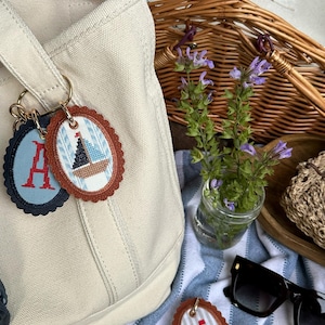 K&ouml;nnte beinhalten: Eine beigefarbene Canvas-Tragetasche mit mehreren ovalen Schl&uuml;sselanh&auml;ngern. Die Schl&uuml;sselanh&auml;nger haben gestickte Designs, darunter ein Segelboot, der Buchstabe "A", eine Ente, eine Golf-Flagge und ein Baseball. Ein Picknickkorb und eine Sonnenbrille sind im Hintergrund.