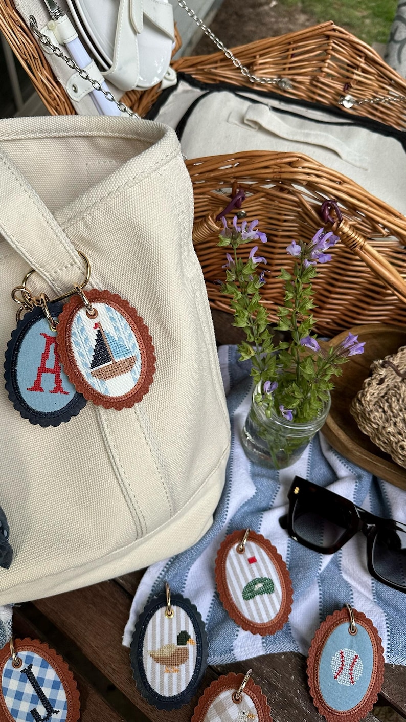 K&ouml;nnte beinhalten: Eine beigefarbene Canvas-Tragetasche mit mehreren ovalen Schl&uuml;sselanh&auml;ngern. Die Schl&uuml;sselanh&auml;nger haben gestickte Designs, darunter ein Segelboot, der Buchstabe "A", eine Ente, eine Golf-Flagge und ein Baseball. Ein Picknickkorb und eine Sonnenbrille sind im Hintergrund.