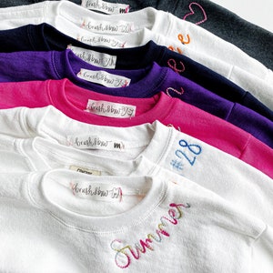 May include: A stack of white, pink, purple, navy blue, and gray sweatshirts with embroidered designs. The designs include the words "Summer", "#28", and "brush & bow".