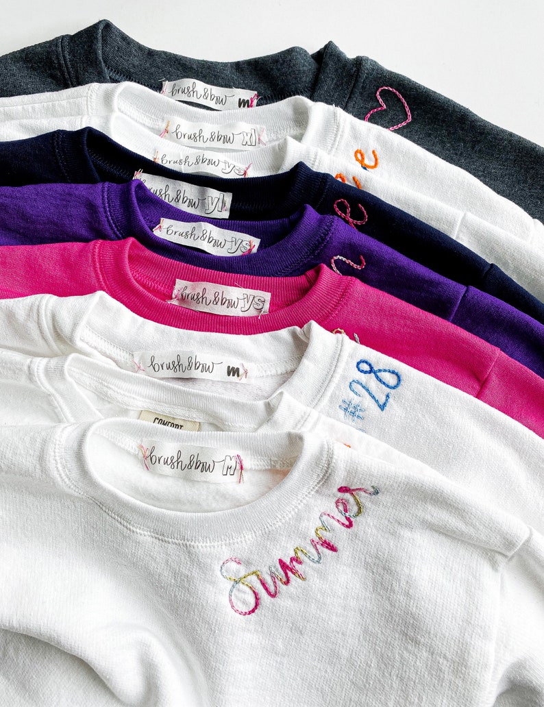 May include: A stack of white, pink, purple, navy blue, and gray sweatshirts with embroidered designs. The designs include the words "Summer", "#28", and "brush & bow".