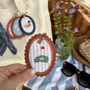 K&ouml;nnte beinhalten: Ovale Schl&uuml;sselanh&auml;nger mit braunem Lederbesatz und goldfarbenen Ringen. Die Schl&uuml;sselanh&auml;nger zeigen gestickte Designs, darunter ein Golfgr&uuml;n, ein Baseball, eine Ente und ein Segelboot. Der Hintergrund umfasst einen Picknickkorb, eine Sonnenbrille und eine Canvas-Tragetasche.