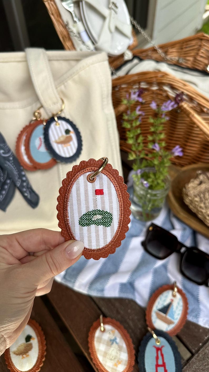 K&ouml;nnte beinhalten: Ovale Schl&uuml;sselanh&auml;nger mit braunem Lederbesatz und goldfarbenen Ringen. Die Schl&uuml;sselanh&auml;nger zeigen gestickte Designs, darunter ein Golfgr&uuml;n, ein Baseball, eine Ente und ein Segelboot. Der Hintergrund umfasst einen Picknickkorb, eine Sonnenbrille und eine Canvas-Tragetasche.