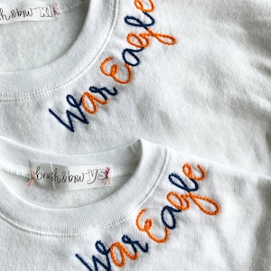 May include: Two white sweatshirts with the words "War Eagle" embroidered on the necklines. The top sweatshirt has the words embroidered in orange and navy blue thread. The bottom sweatshirt has the words embroidered in navy blue and orange thread.