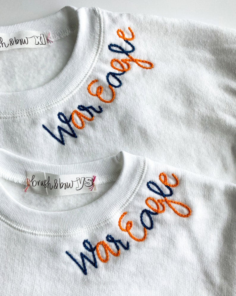 May include: Two white sweatshirts with the words "War Eagle" embroidered on the necklines. The top sweatshirt has the words embroidered in orange and navy blue thread. The bottom sweatshirt has the words embroidered in navy blue and orange thread.