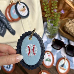 K&ouml;nnte beinhalten: Ovale Schl&uuml;sselanh&auml;nger mit verschiedenen Designs, darunter ein Baseball, ein Segelboot und eine Ente. Der Baseball-Schl&uuml;sselanh&auml;nger ist hellblau mit einem roten Baseball-Naht-Design. Andere Schl&uuml;sselanh&auml;nger haben braune, gewellte R&auml;nder.