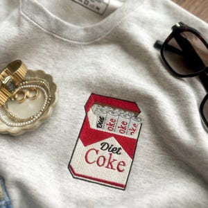 Op de afbeelding: Een lichtgrijs sweatshirt met een geborduurd Diet Coke-pakje. Het rood-witte pakje heeft de tekst "Diet Coke". Het sweatshirt heeft ook een label met "Brush & Dye".