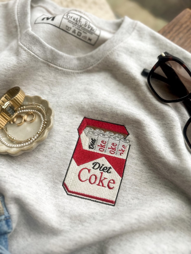 Op de afbeelding: Een lichtgrijs sweatshirt met een geborduurd Diet Coke-pakje. Het rood-witte pakje heeft de tekst "Diet Coke". Het sweatshirt heeft ook een label met "Brush & Dye".
