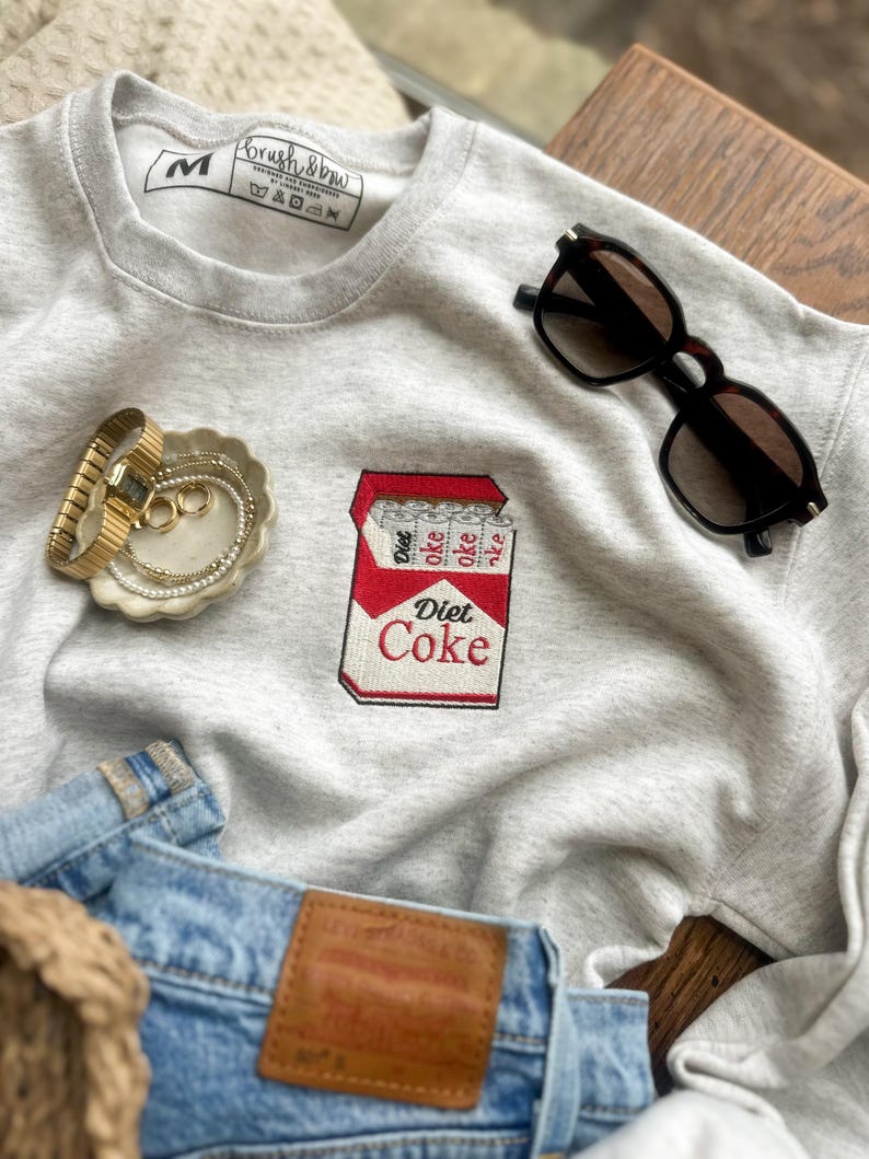 Op de afbeelding: Een grijze sweatshirt met een Diet Coke-ontwerp. Het rood-witte geborduurde ontwerp toont een Diet Coke-pakje. Zonnebril, een gouden horloge en sieraden zijn ook in de afbeelding te zien.