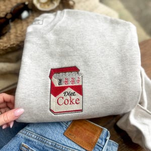 Op de afbeelding: Een lichtgrijs sweatshirt met een geborduurd Diet Coke-ontwerp in rood en wit. Het sweatshirt is opgevouwen bovenop een blauwe spijkerbroek. De jeans heeft een bruin leren label op de achterzak.