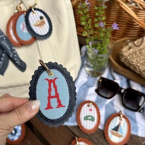 K&ouml;nnte beinhalten: Ovale Schl&uuml;sselanh&auml;nger mit verschiedenen gestickten Designs, darunter ein rotes "A", ein Baseball, eine Ente und ein Segelboot. Die Schl&uuml;sselanh&auml;nger haben eine braune Lederunterseite und einen blau-marinefarbenen, gewellten Rand. Eine beigefarbene Tragetasche und ein Picknickkorb sind im Hintergrund.