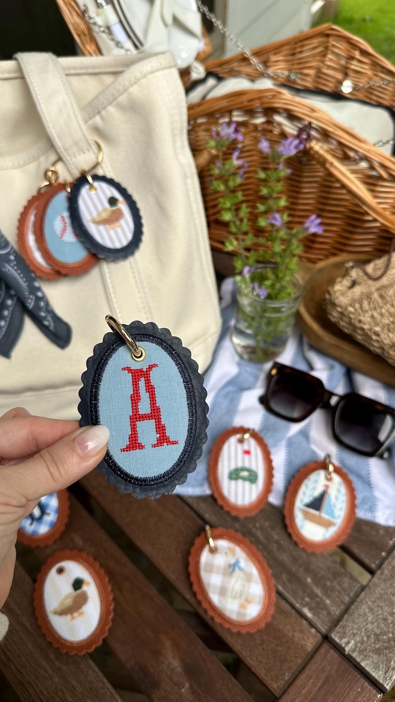 K&ouml;nnte beinhalten: Ovale Schl&uuml;sselanh&auml;nger mit verschiedenen gestickten Designs, darunter ein rotes "A", ein Baseball, eine Ente und ein Segelboot. Die Schl&uuml;sselanh&auml;nger haben eine braune Lederunterseite und einen blau-marinefarbenen, gewellten Rand. Eine beigefarbene Tragetasche und ein Picknickkorb sind im Hintergrund.