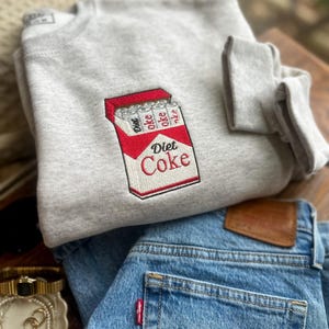 Op de afbeelding: Een lichtgrijs sweatshirt met een geborduurd Diet Coke-pakje. Het ontwerp toont een rood en wit pakje met de woorden "Diet Coke" in het rood. Daaronder is een opgevouwen blauwe spijkerbroek te zien, met een klein rood label.