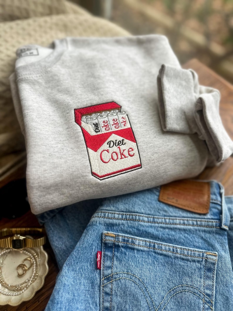 Op de afbeelding: Een lichtgrijs sweatshirt met een geborduurd Diet Coke-pakje. Het ontwerp toont een rood en wit pakje met de woorden "Diet Coke" in het rood. Daaronder is een opgevouwen blauwe spijkerbroek te zien, met een klein rood label.