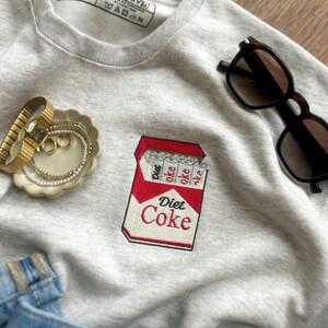 Op de afbeelding: Een lichtgrijs sweatshirt met een geborduurd Diet Coke-ontwerp. De sweatshirt heeft een ronde hals en een klein label bij de halslijn. Zonnebril en sieraden zijn ook zichtbaar.