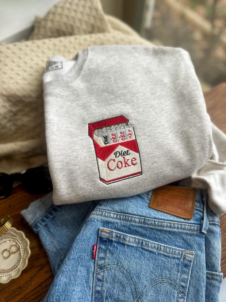 Op de afbeelding: Een lichtgrijs sweatshirt met een geborduurd Diet Coke-pakketontwerp. Het sweatshirt wordt gecombineerd met lichtblauwe denim jeans. Een goudkleurige armband en zonnebril zijn ook zichtbaar.