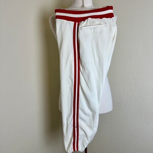 May include: White baseball pants with a red stripe down the side and red waistband. The pants are displayed on a mannequin. The pants have a button and a pocket on the back.