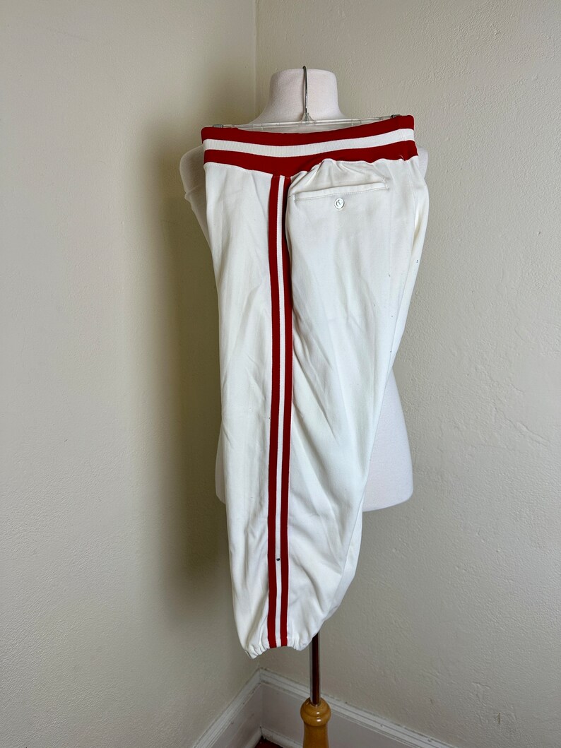 May include: White baseball pants with a red stripe down the side and red waistband. The pants are displayed on a mannequin. The pants have a button and a pocket on the back.