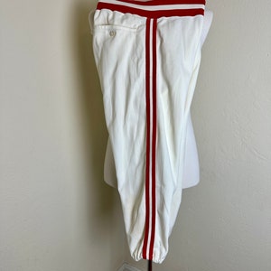May include: Cream-colored athletic pants with a red stripe down each side and a red band at the waist. The pants are displayed on a mannequin. The pants have a button closure and a pocket on the back.