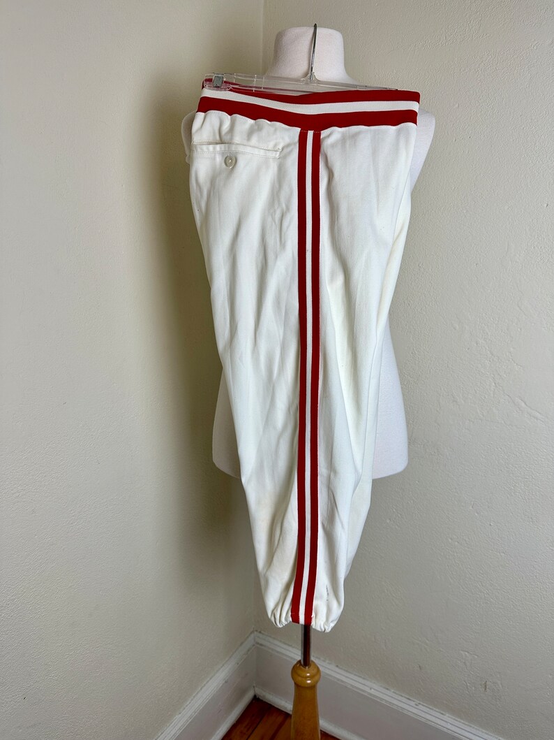 May include: Cream-colored athletic pants with a red stripe down each side and a red band at the waist. The pants are displayed on a mannequin. The pants have a button closure and a pocket on the back.