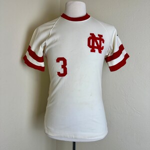 May include: Cream-colored vintage athletic jersey with red accents. Features a red "NC" logo on the chest and the number "3" below. The sleeves have red stripes, and the collar is also red. The jersey is displayed on a mannequin.