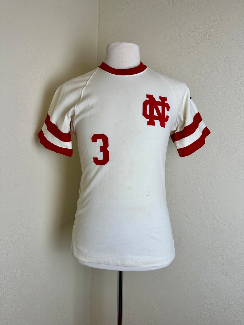 May include: Cream-colored vintage athletic jersey with red accents. Features a red "NC" logo on the chest and the number "3" below. The sleeves have red stripes, and the collar is also red. The jersey is displayed on a mannequin.