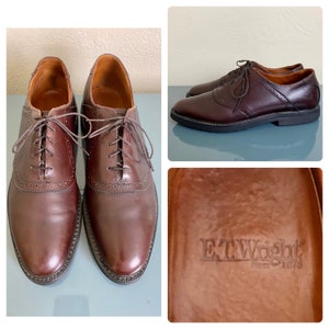 May include: A pair of brown leather dress shoes with a lace-up design. The shoes have a classic, formal style with decorative perforations along the seams. The soles are dark brown with a black border. The brand name "E.T. Wright" is visible.