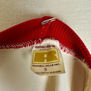 May include: Close-up of a vintage athletic shirt collar. The collar is red with a white trim and a metal clasp. A tag reads "RUSSELL ATHLETIC DIVISION OF" and "RUSSELL MILLS INC." with a size "S".