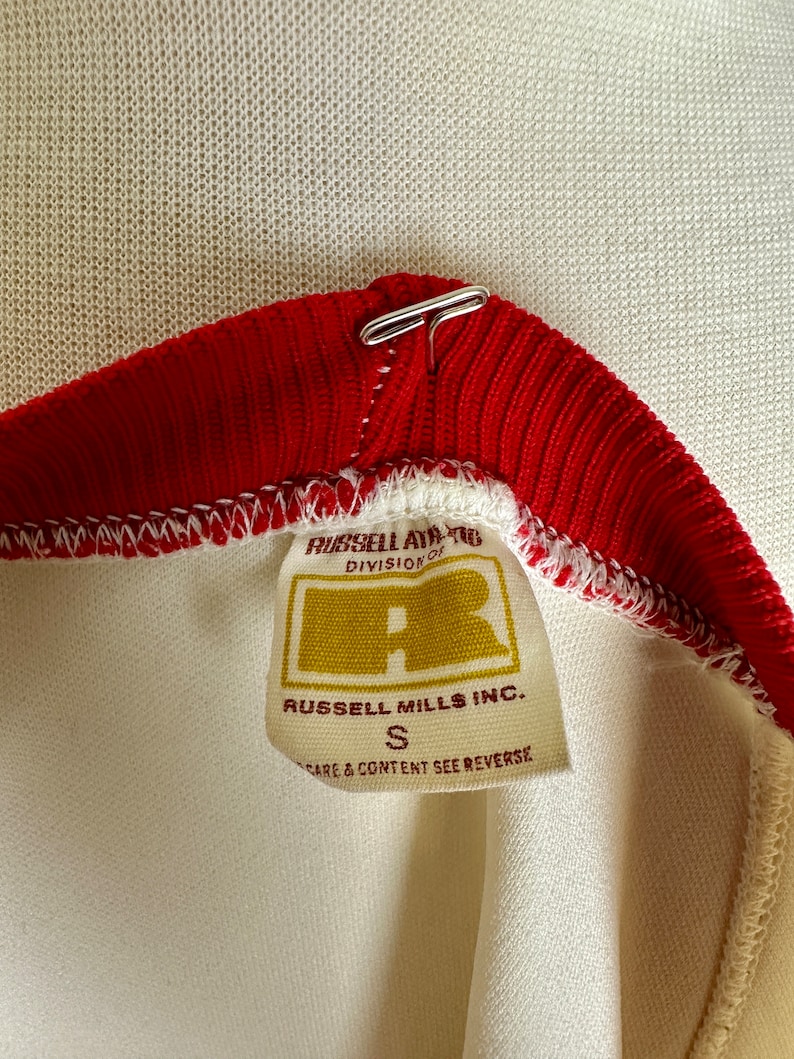 May include: Close-up of a vintage athletic shirt collar. The collar is red with a white trim and a metal clasp. A tag reads "RUSSELL ATHLETIC DIVISION OF" and "RUSSELL MILLS INC." with a size "S".