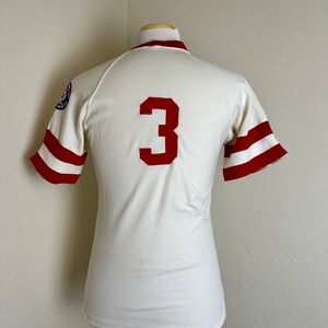 May include: A vintage baseball jersey in cream with red accents. The jersey features a large red number "3" on the back and red stripes on the sleeves. A patch is visible on the left sleeve. The jersey is displayed on a mannequin.