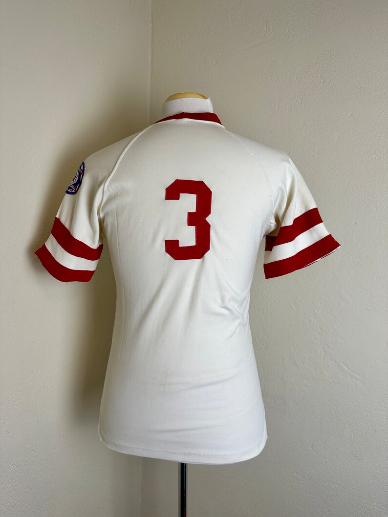May include: A vintage baseball jersey in cream with red accents. The jersey features a large red number "3" on the back and red stripes on the sleeves. A patch is visible on the left sleeve. The jersey is displayed on a mannequin.