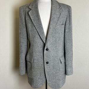 May include: A gray tweed blazer displayed on a mannequin. The jacket has a notched lapel, two front pockets, and two black buttons. The fabric has a speckled texture, and the blazer is a classic, tailored style.