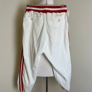 May include: White baseball pants with red stripes down the sides and a red waistband. The pants have two back pockets and are displayed on a mannequin. The pants are made of a light-colored fabric.