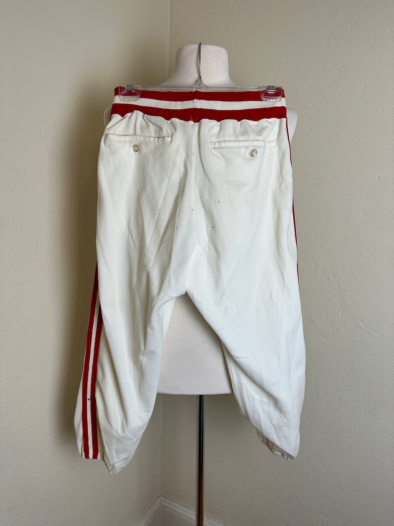 May include: White baseball pants with red stripes down the sides and a red waistband. The pants have two back pockets and are displayed on a mannequin. The pants are made of a light-colored fabric.