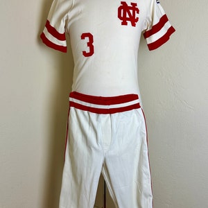 May include: Vintage baseball uniform in cream with red accents. The jersey features the number 3 and the letters "NC" in red. The uniform includes a short-sleeved shirt and matching pants.