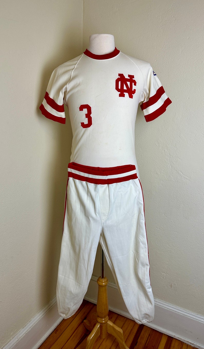 May include: Vintage baseball uniform in cream with red accents. The jersey features the number 3 and the letters "NC" in red. The uniform includes a short-sleeved shirt and matching pants.