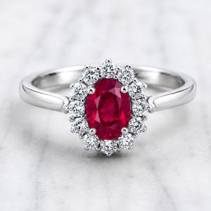 May include: A silver-toned ring featuring a central oval-cut deep red gemstone surrounded by a halo of twelve small, round, clear white stones. The band is smooth and polished, connecting to the gemstone setting with a simple, clean design.