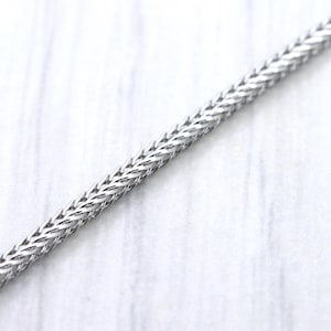 May include: Close-up of a silver chain with a woven design. The chain is set against a white, textured background. The chain's links are tightly woven, creating a textured surface.