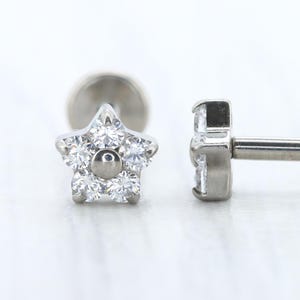 May include: Two silver-colored, star-shaped earrings with clear, sparkling stones. The earrings have a flat back and a post for piercing. The star design is made of multiple small stones, and the center has a single, larger stone.