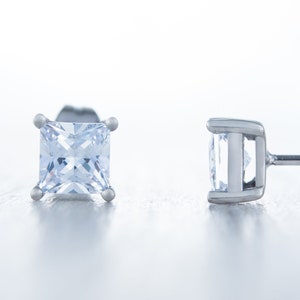 May include: A pair of square-cut, light blue gemstone stud earrings. The earrings are set in a silver metal, with a post and butterfly backing. The gemstones are clear and faceted, reflecting light. The earrings are displayed on a light, reflective surface.