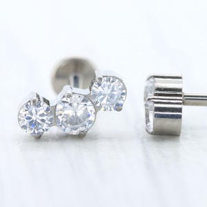 May include: Two silver labret earrings. One has three clear, round, sparkling stones clustered together. The other has a single, clear, round, sparkling stone. Both earrings have a flat back and a post for piercing.